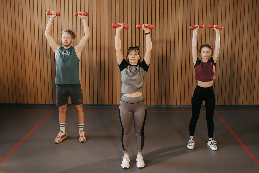 Three adults press dumbbells overhead in a strength class