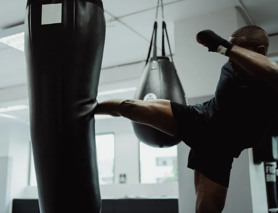 A man in black training gear throws a roundhouse kick into a heavy bag in a bright studio